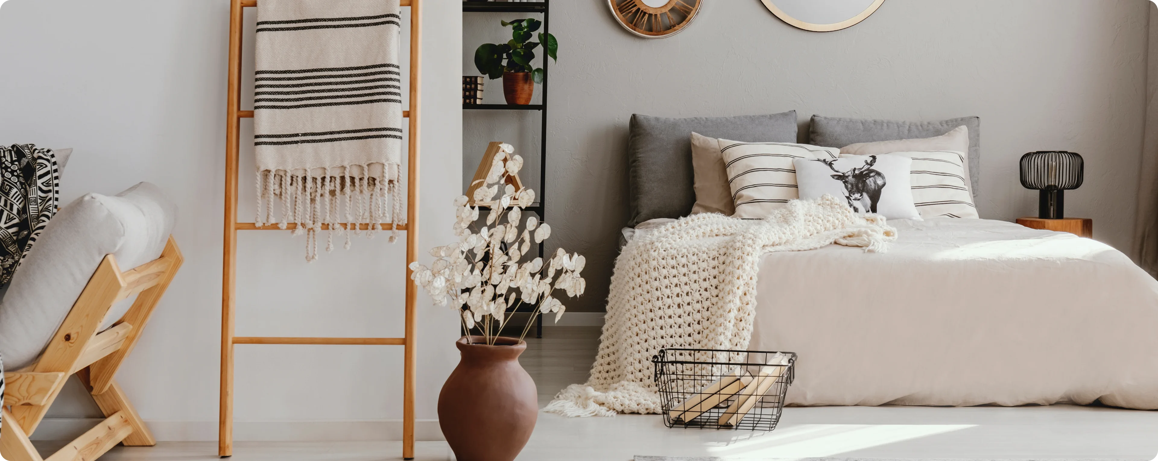 Cozy bedroom with a bed, gray pillows, beige blanket, decorative ladder holding a striped towel, potted plant, and a round vase with flowers. Wall mirrors and a shelf are in the background.