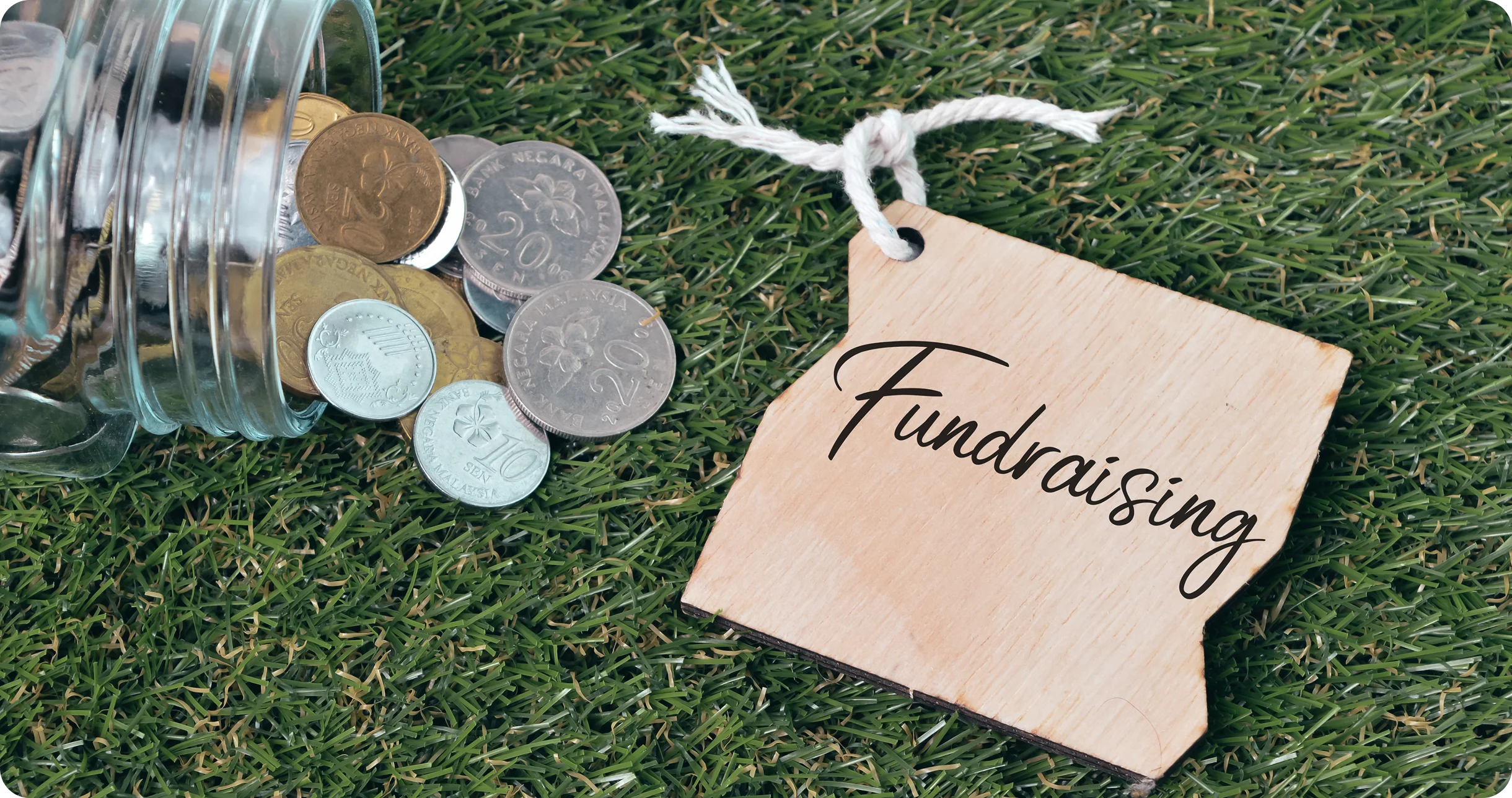Coins spill from a jar onto grass next to a wooden tag with "Fundraising" written on it.